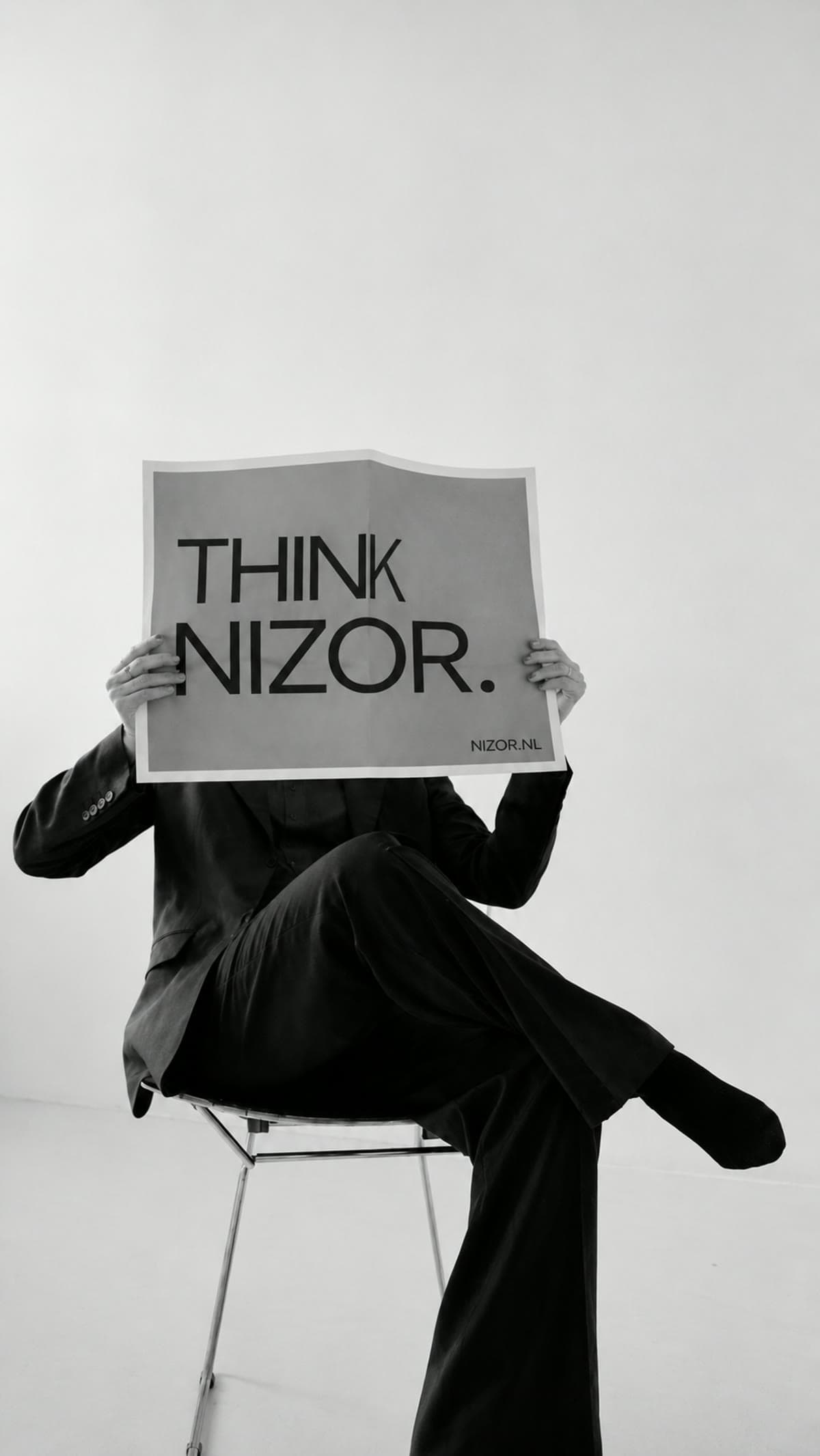Iemand in pak op een stoel, met een poster voor het gezicht waarop THINK NIZOR staat